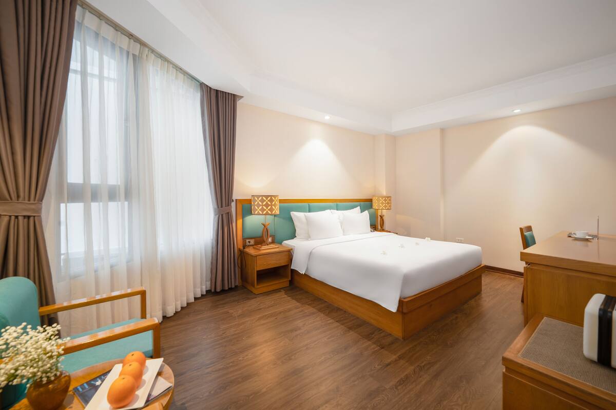 Habitación de hotel con cama, mesitas de noche de madera, escritorio, silla y ventana con cortinas.