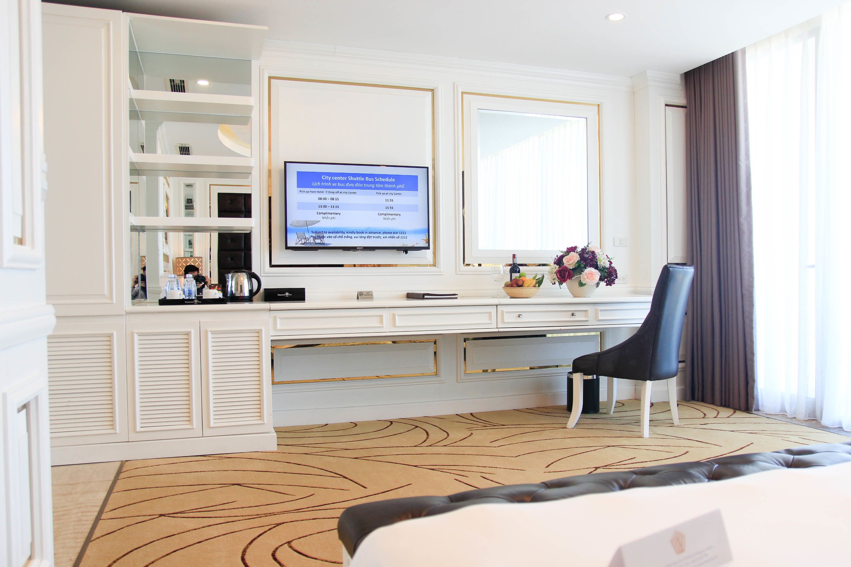 A hotel room with a large flat-screen TV displaying a shuttle bus schedule, a black leather chair, a white desk with drawers, and a patterned carpet.