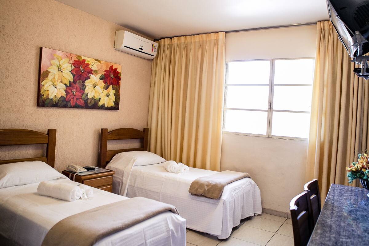 Een hotelkamer met twee bedden, een schilderij aan de muur, een airconditioner, een raam met gordijnen en een bureau met stoel.