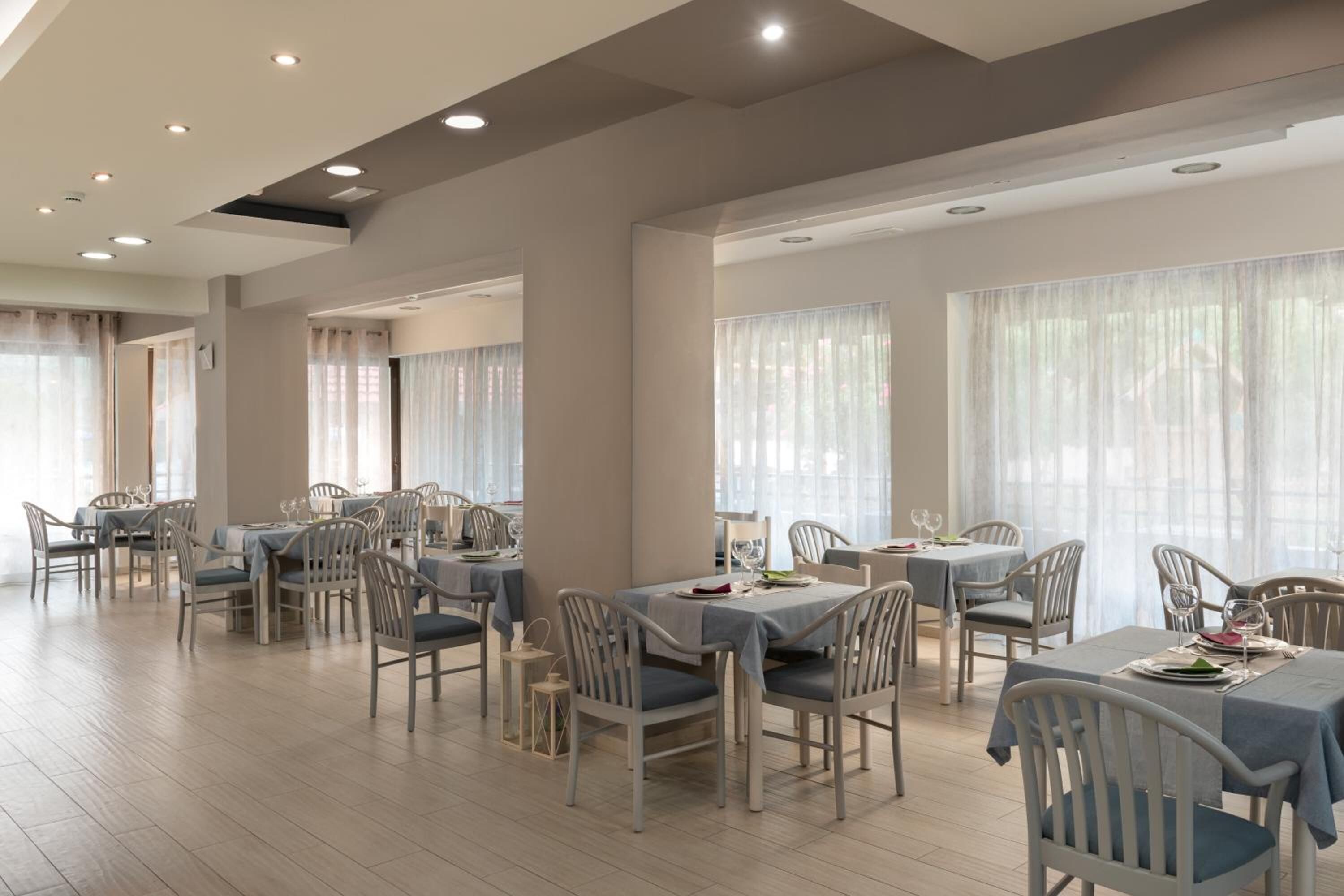 A spacious dining area with multiple tables and chairs, large windows with sheer curtains, and recessed ceiling lighting.