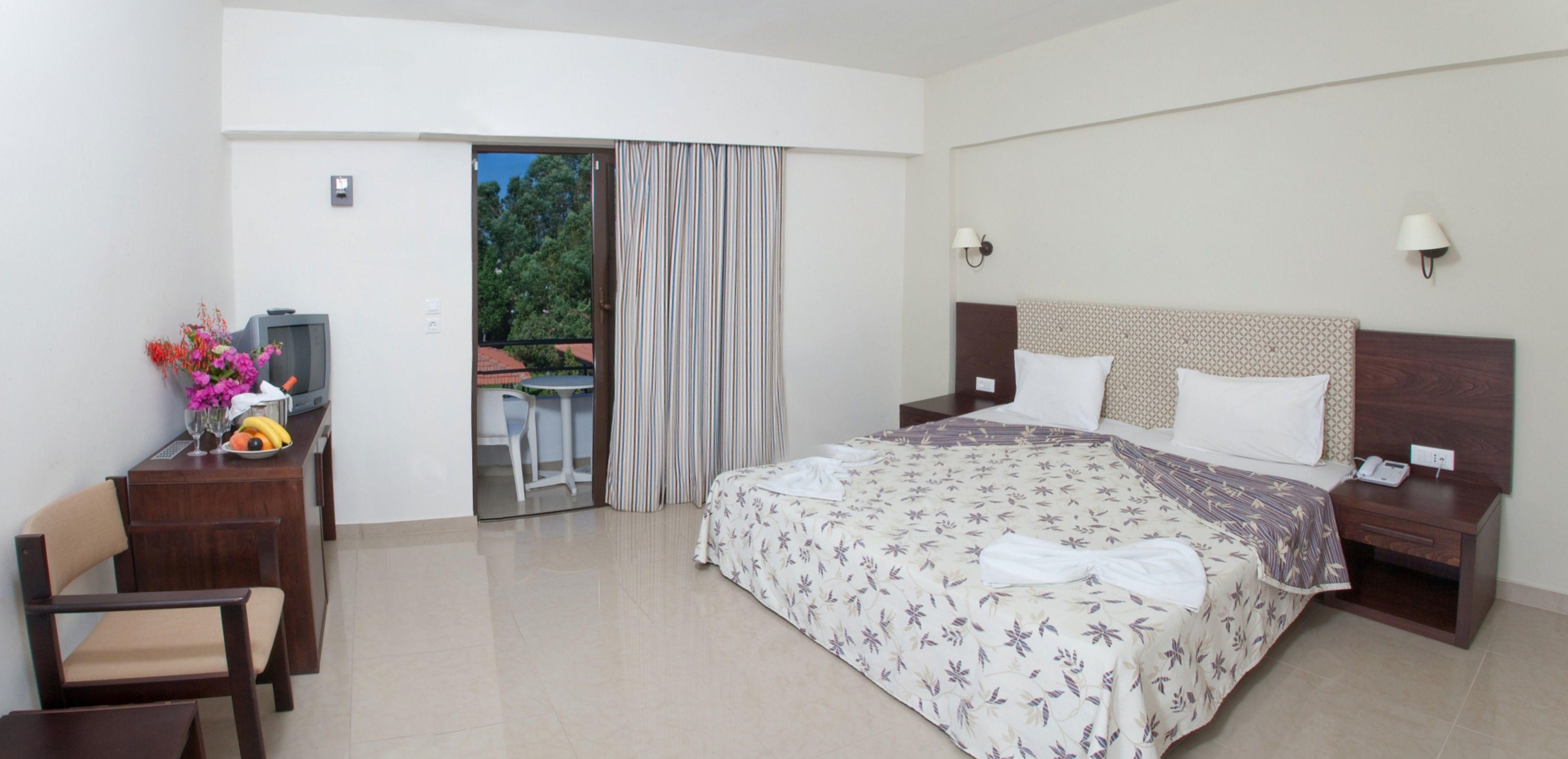 A hotel room with a bed, a desk with a chair, a TV, and a balcony with curtains.