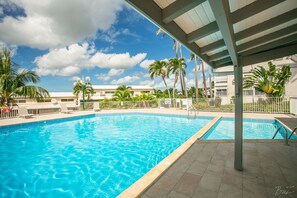 A clear blue pool with a tiled deck, surrounded by palm trees and a covered area with a metal roof.