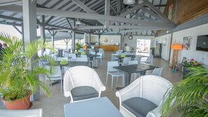 A spacious outdoor cafe with white wicker furniture, potted plants, and a central bar area. - Hotel Village Soleil (Le Gosier)