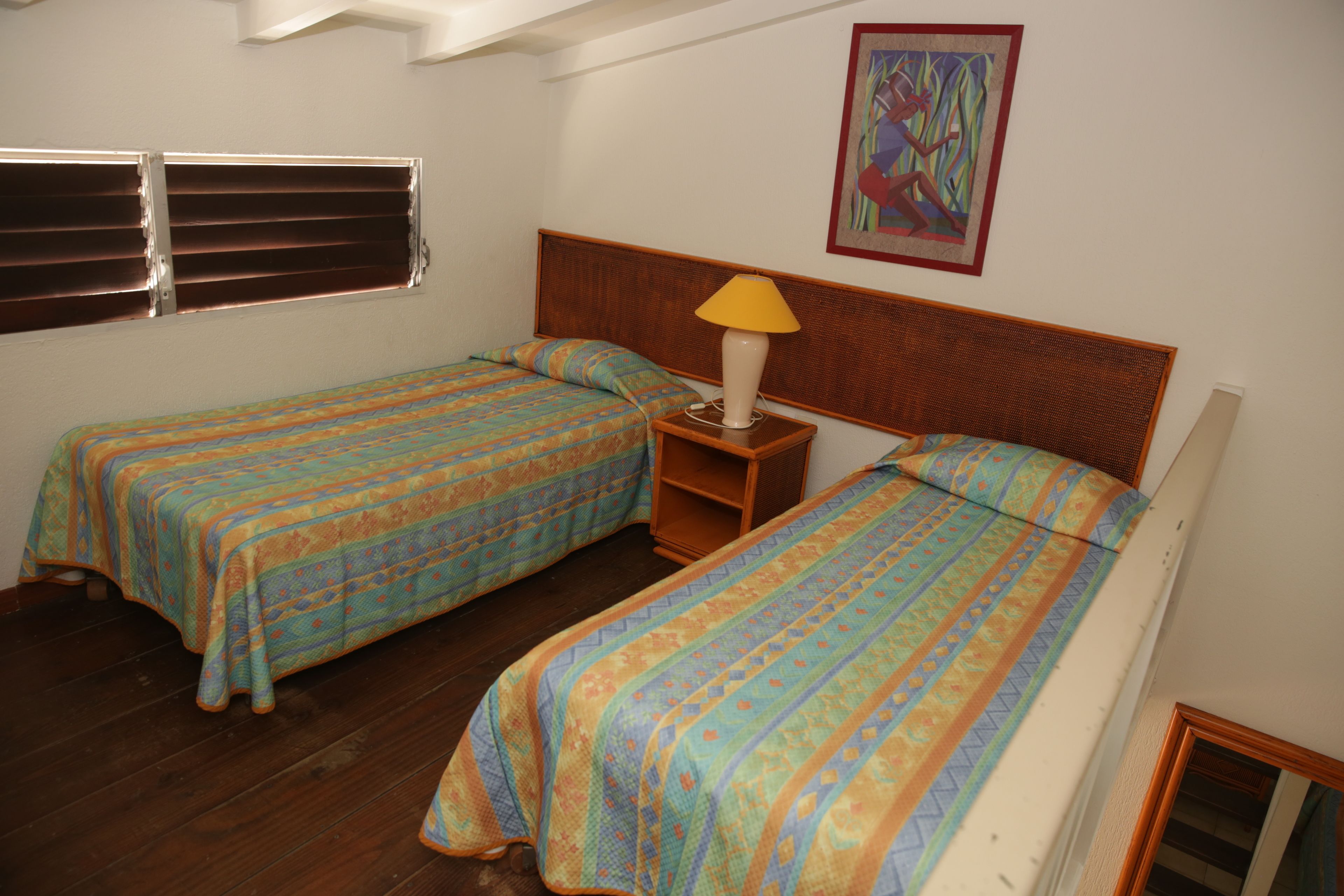 A room with two single beds, a wooden headboard, a bedside table with a lamp, and a window with blinds.