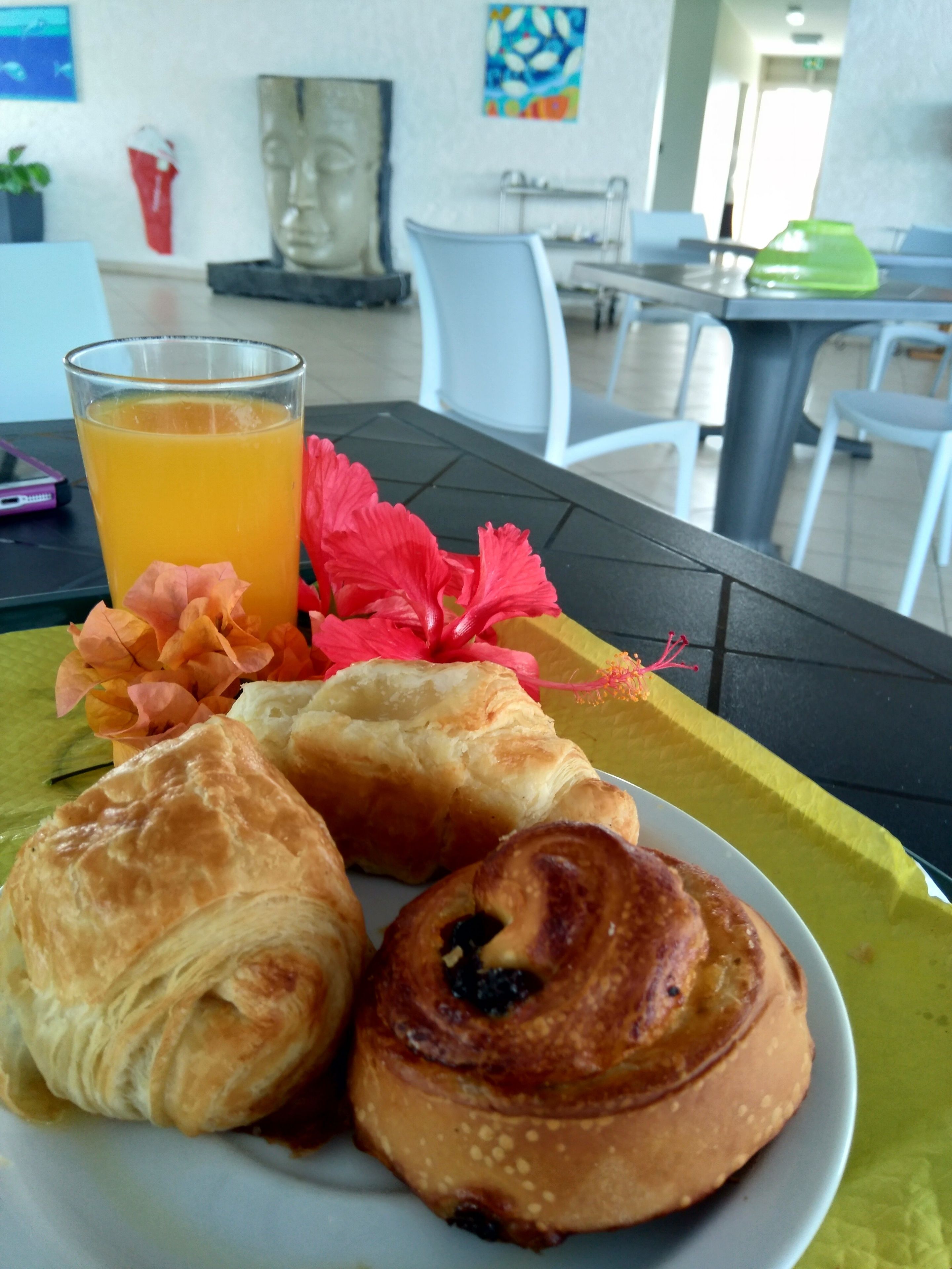 Une table mise pour le petit-déjeuner, avec des croissants et un verre de jus d’orange, un vase de fleurs et un tableau en arrière-plan.