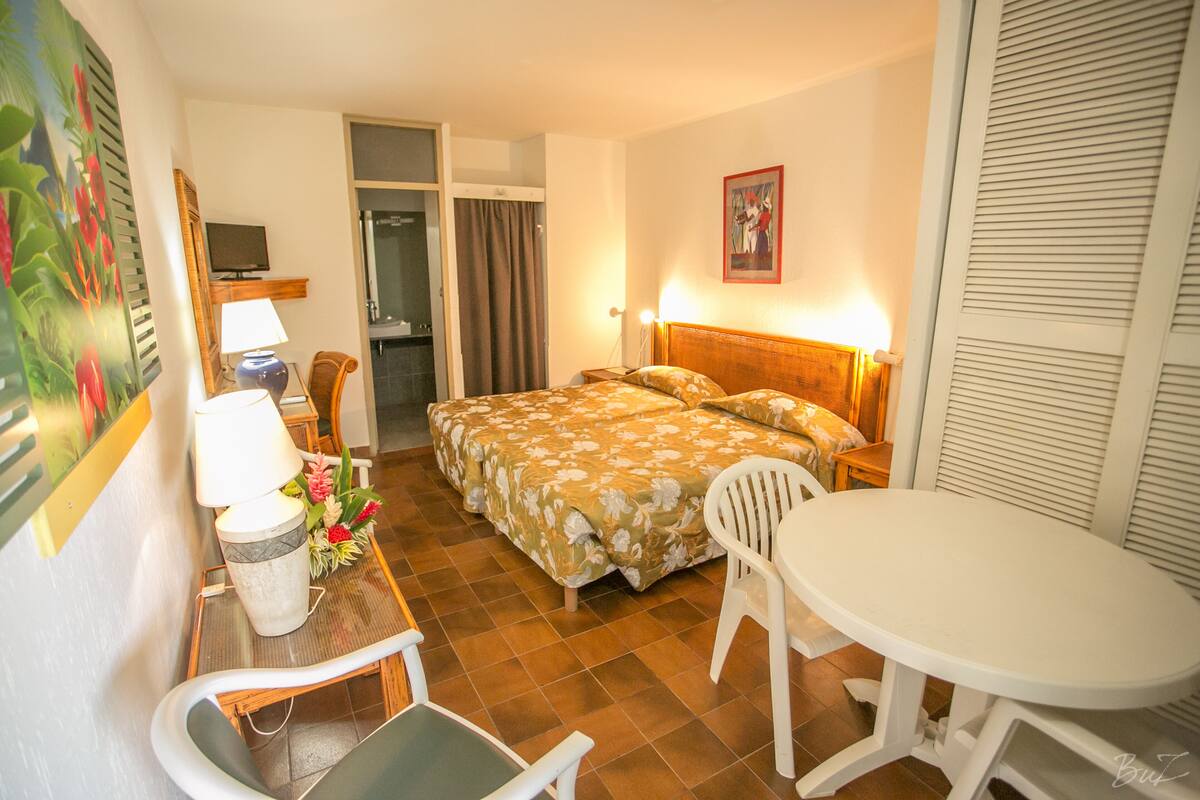 A hotel room with a bed, a desk with a lamp, a chair, a small table, and a shower area.