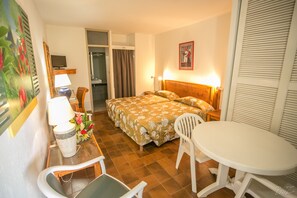 A hotel room with a bed, a desk with a lamp, a chair, a small table, and a shower area.