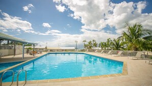 A swimming pool with a tiled deck, lounge chairs, and palm trees. - Hotel Village Soleil (Le Gosier)
