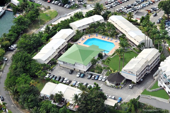 An aerial view of a hotel with a swimming pool, surrounded by lush greenery and parked cars.