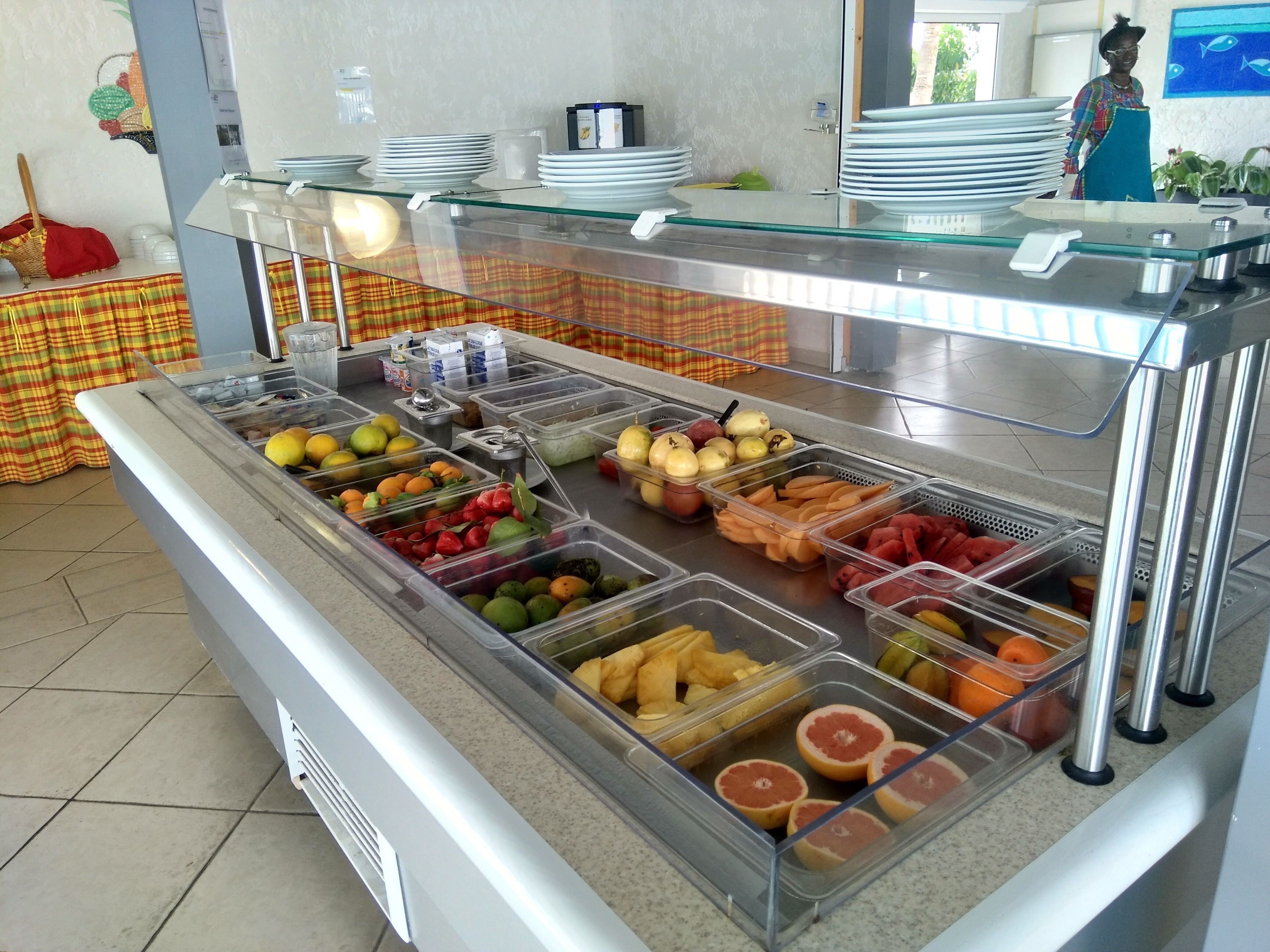 A buffet area with various fruits and vegetables in clear containers, a stack of plates, and a person in the background.