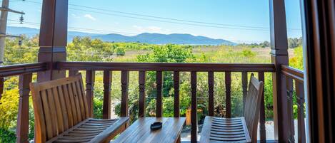 Un balcón de madera con dos sillas y una mesa, con vistas a un paisaje verde y montañas al fondo.