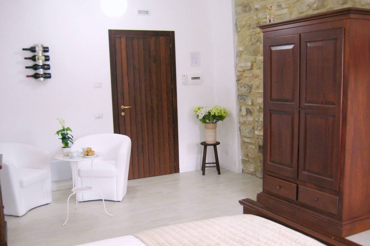 A hotel room with a bed, a white armchair, a small table with a plant, and a wooden wardrobe.