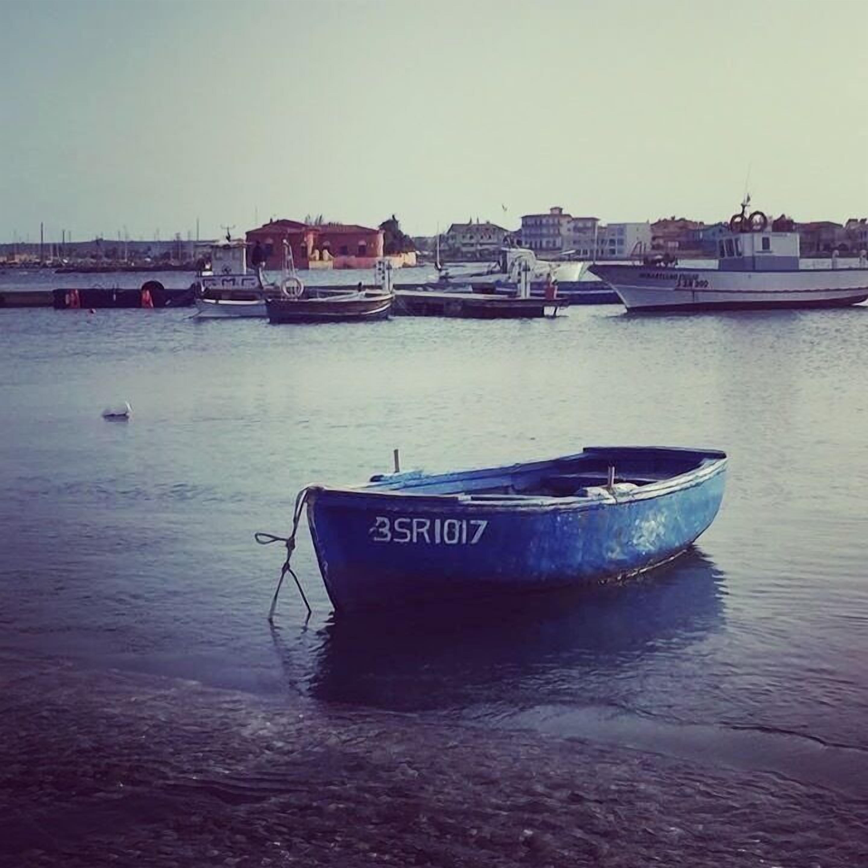 Um porto tranquilo com várias embarcações atracadas, um barco azul em destaque com o número 3SR1017 em primeiro plano, e edifícios ao fundo.
