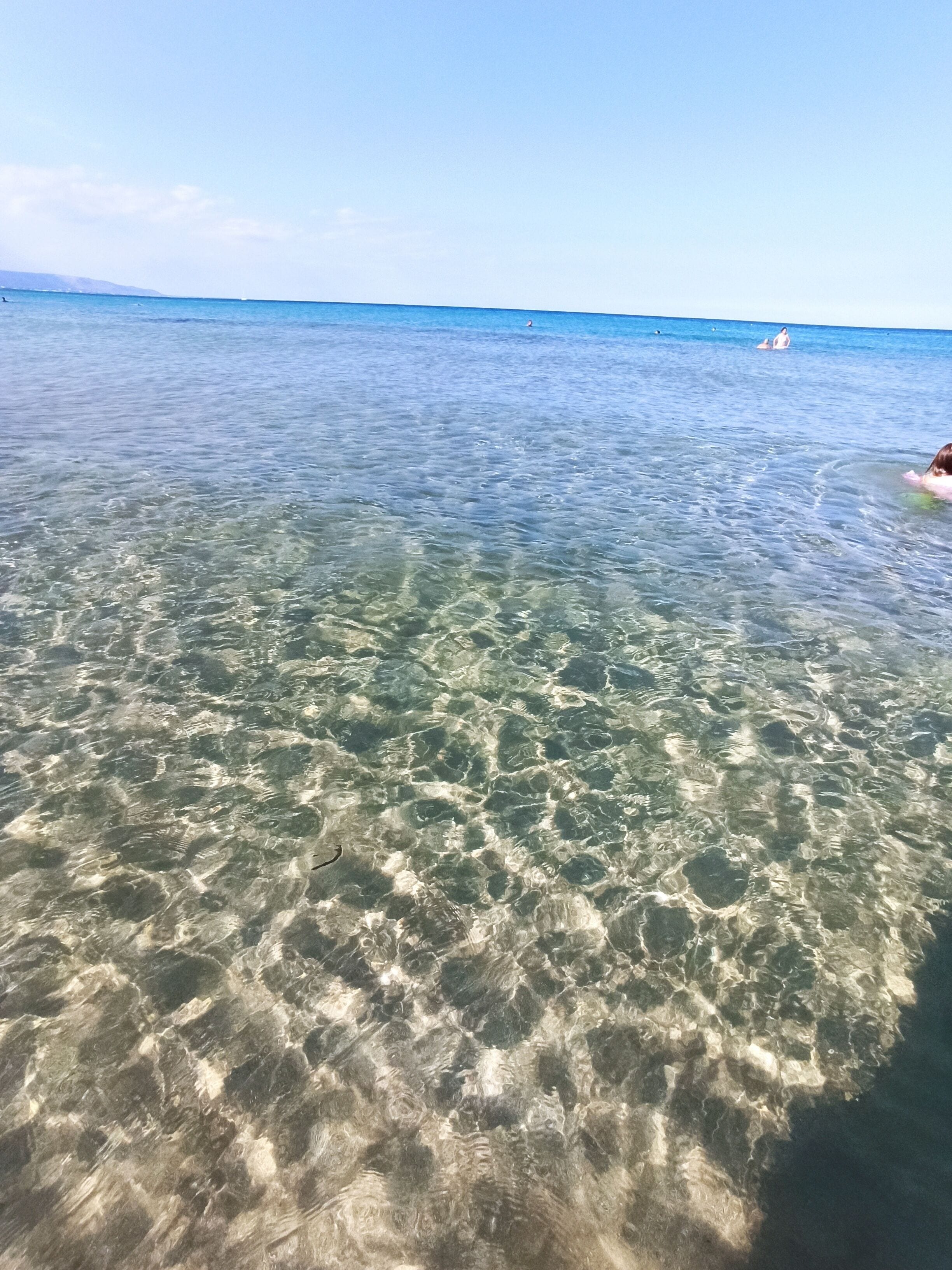 Agua poco profunda y transparente con patrones visibles bajo la superficie, un horizonte lejano y una persona en el agua.