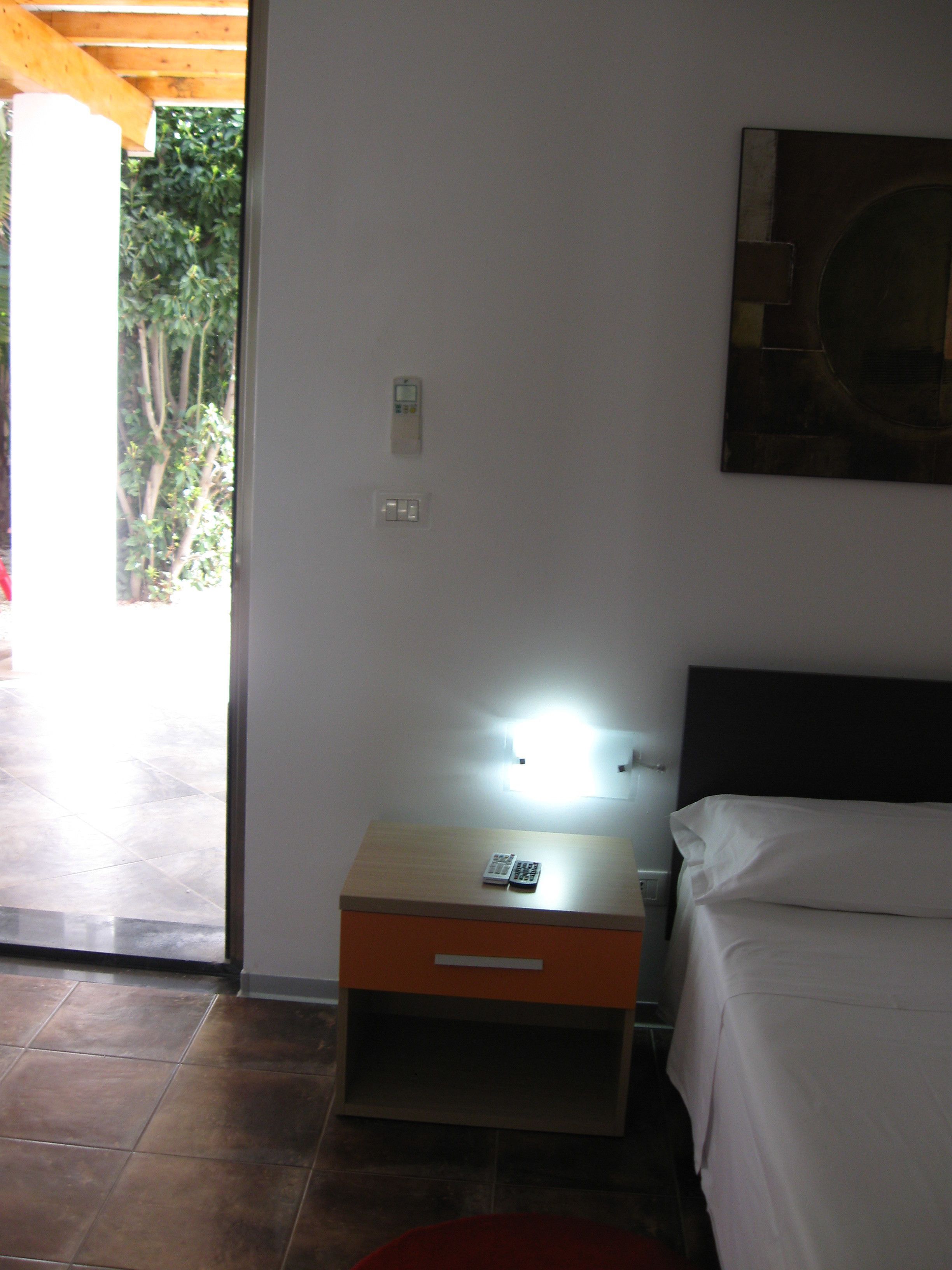 A hotel room with a bed, a nightstand with a lamp, a remote control, and a wall-mounted light switch.