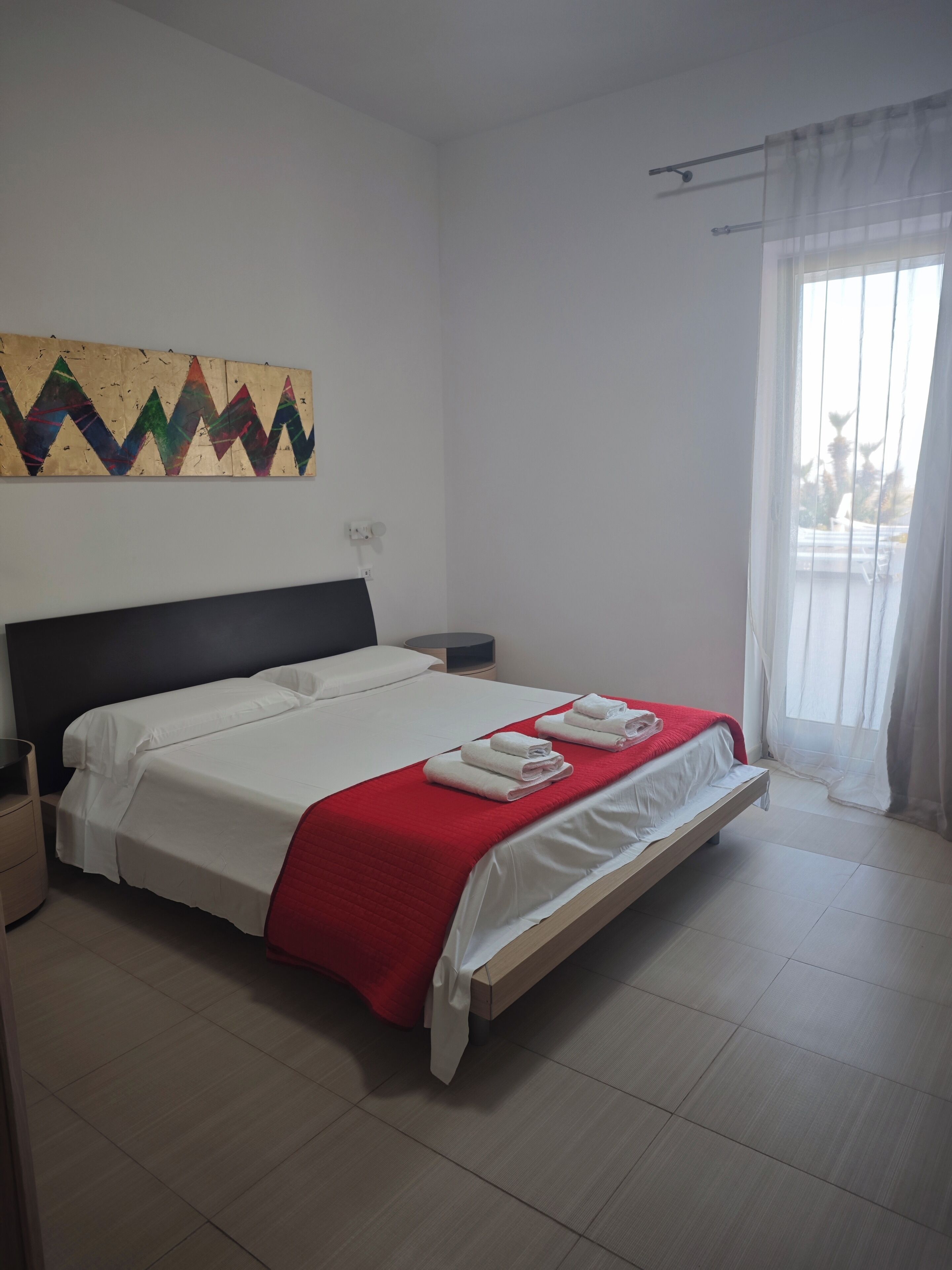 A neatly made bed with white linens and a red blanket, a wall art piece, and a window with sheer curtains.