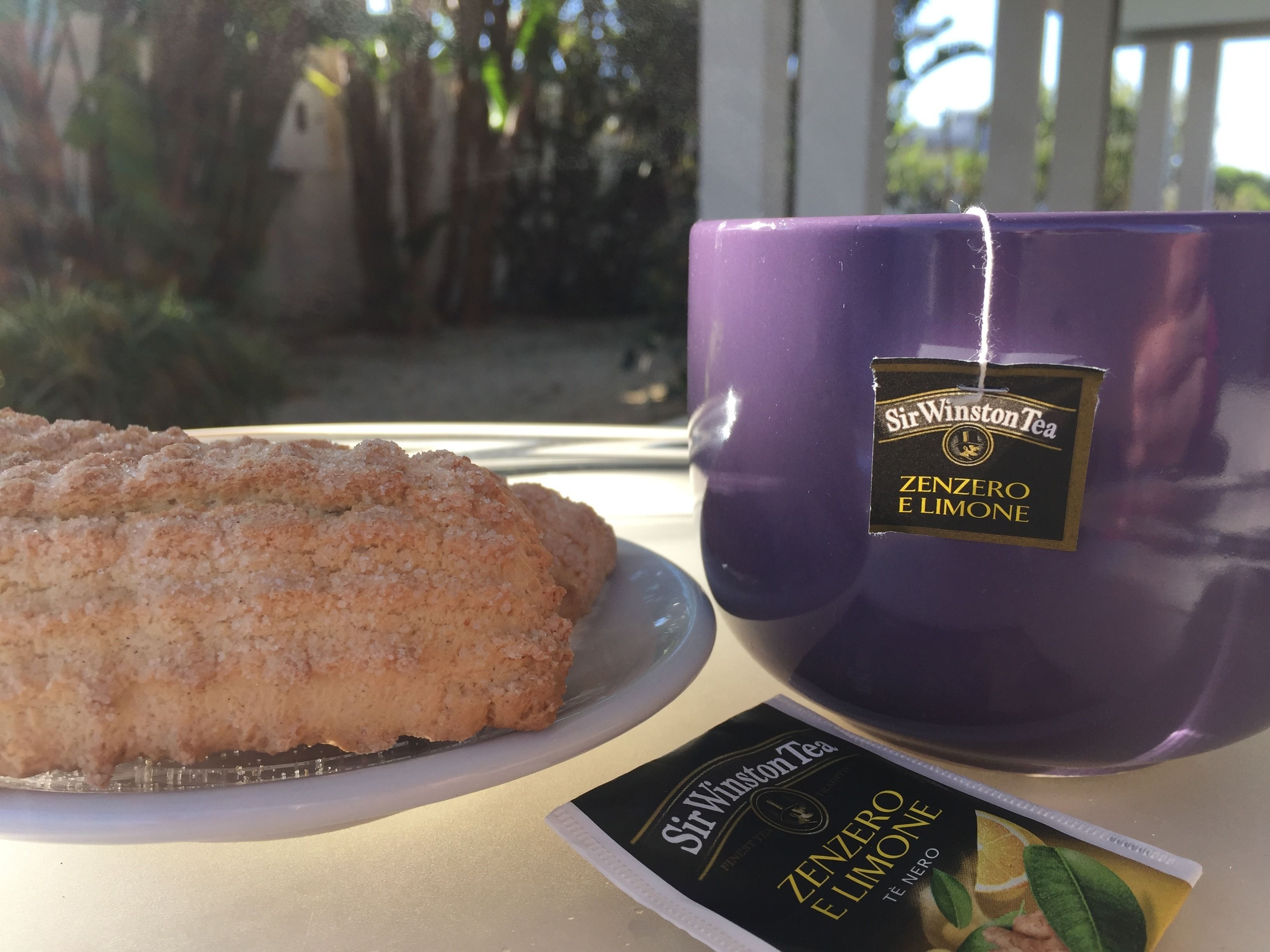 Una bustina di tè Sir Winston Tea Zenzero e Limone viola, un piatto con una pasticceria e una confezione di tè su un tavolo.