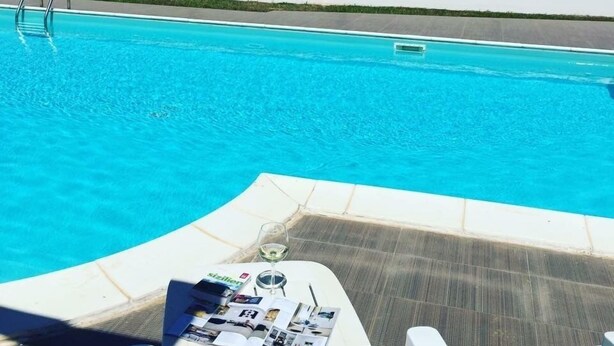 A pool with clear blue water, a lounge area with a magazine, and a view of the ocean and distant landscape. - B&B Resort Torrefano (Pachino)