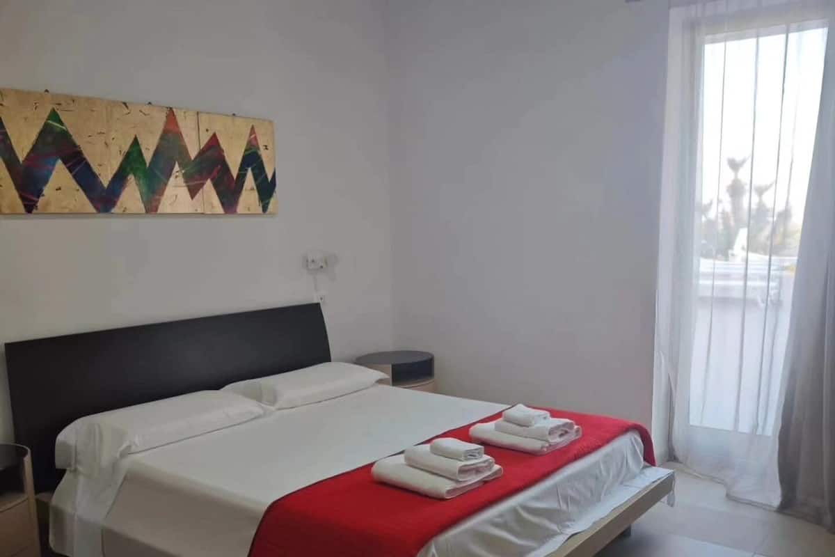Habitación de hotel con cama, mesita de noche, una manta roja y un cuadro abstracto colorido en la pared.