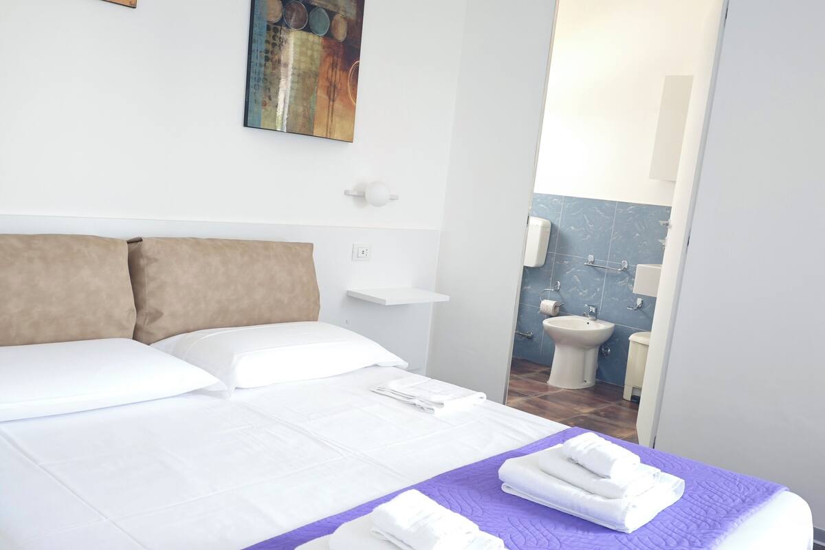 Habitación de hotel con una cama grande, un baño visible a través de una puerta abierta y obras de arte en las paredes.