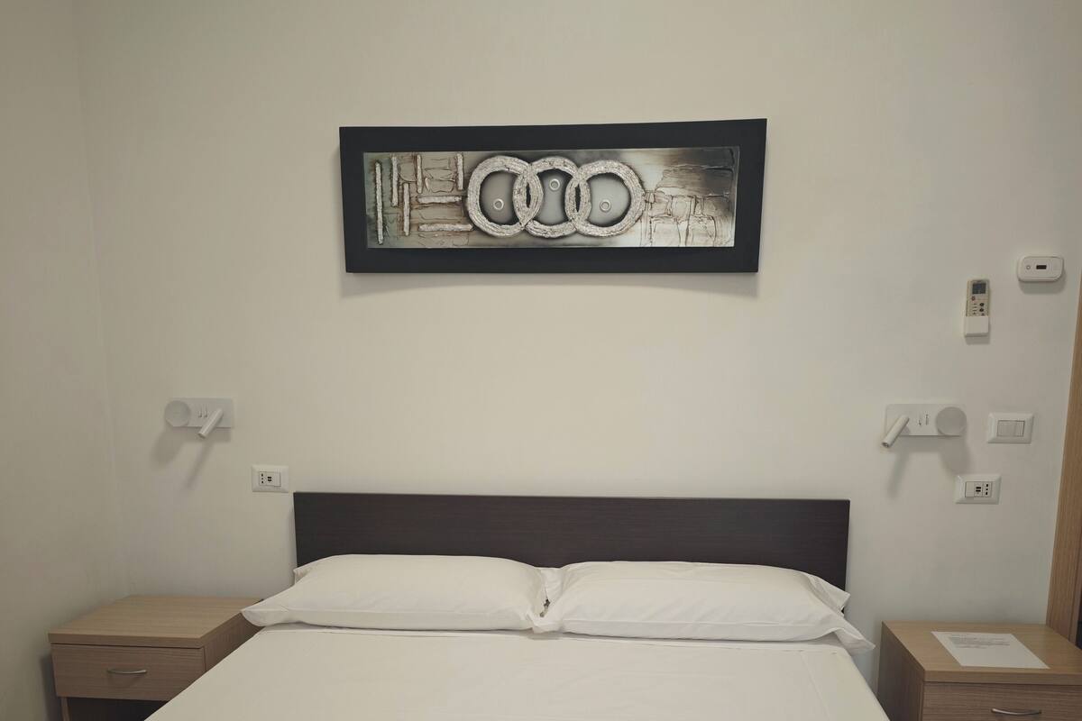 Habitación de hotel con cama, dos mesitas de noche, una obra de arte colgada en la pared y un panel de control en la pared.
