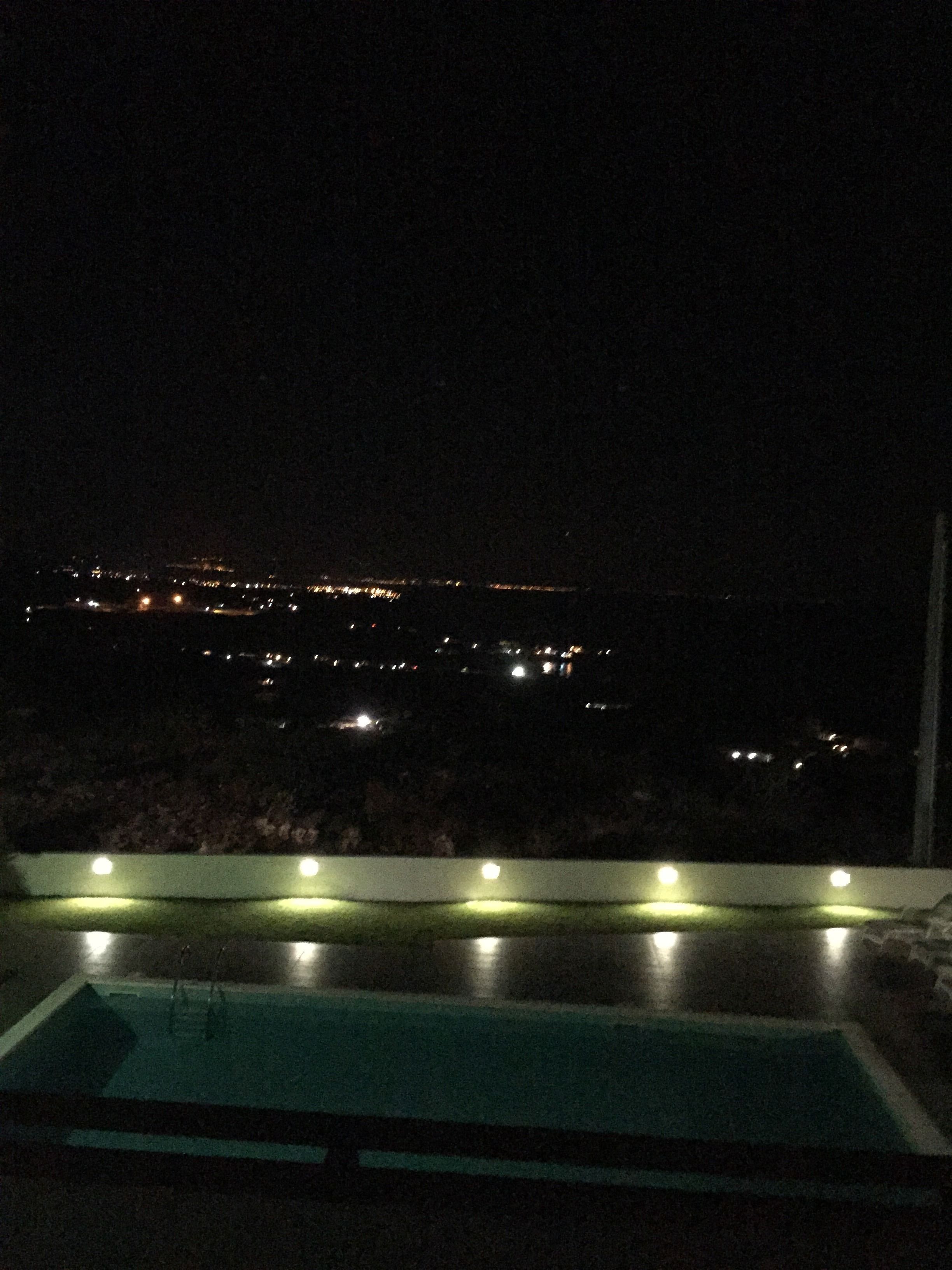 A nighttime view from a balcony with a pool, city lights in the distance, and a railing with lights.