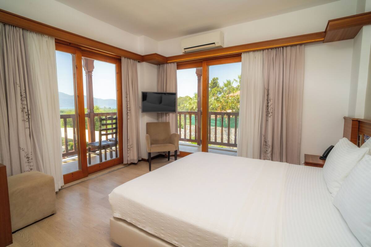 A hotel room with a large bed, a chair, a TV, and a balcony with a view of greenery and mountains.