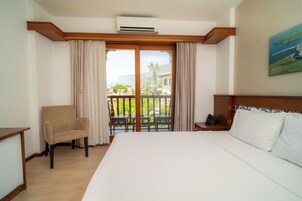 Una habitación de hotel con cama, una silla, un escritorio y un balcón con vistas a zonas verdes y edificios.