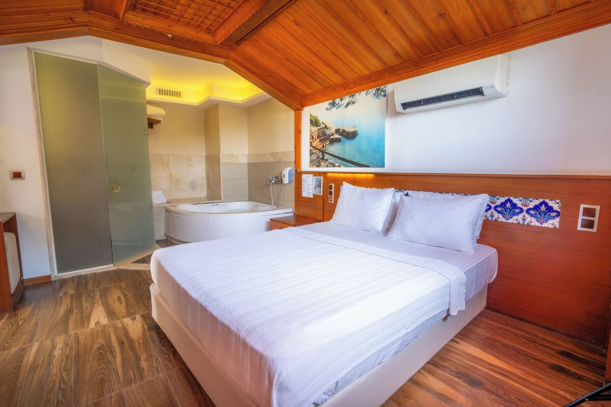 A bedroom with a large bed, a wooden ceiling, a bathroom with a bathtub, and a painting on the wall.