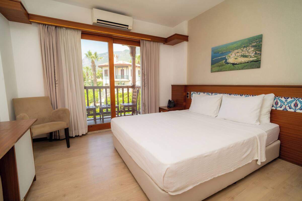 A hotel room with a bed, a chair, a desk, and a balcony with a view of a coastal area.