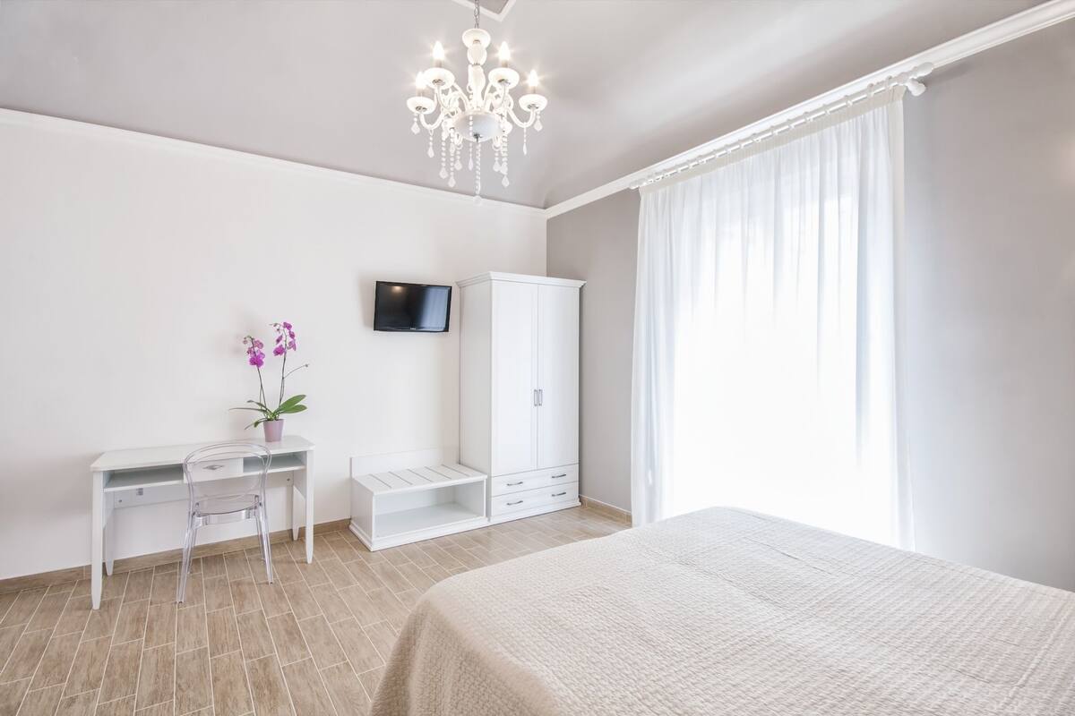 A bedroom with a bed, a desk with a chair, a wardrobe, a television on the wall, and a window with curtains.