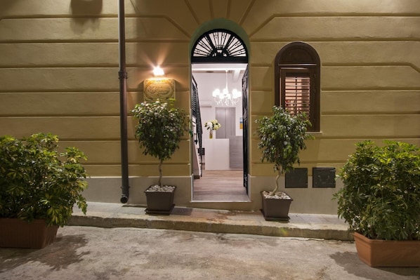 A building entrance with a covered walkway, potted plants, and a visible interior through an open door.