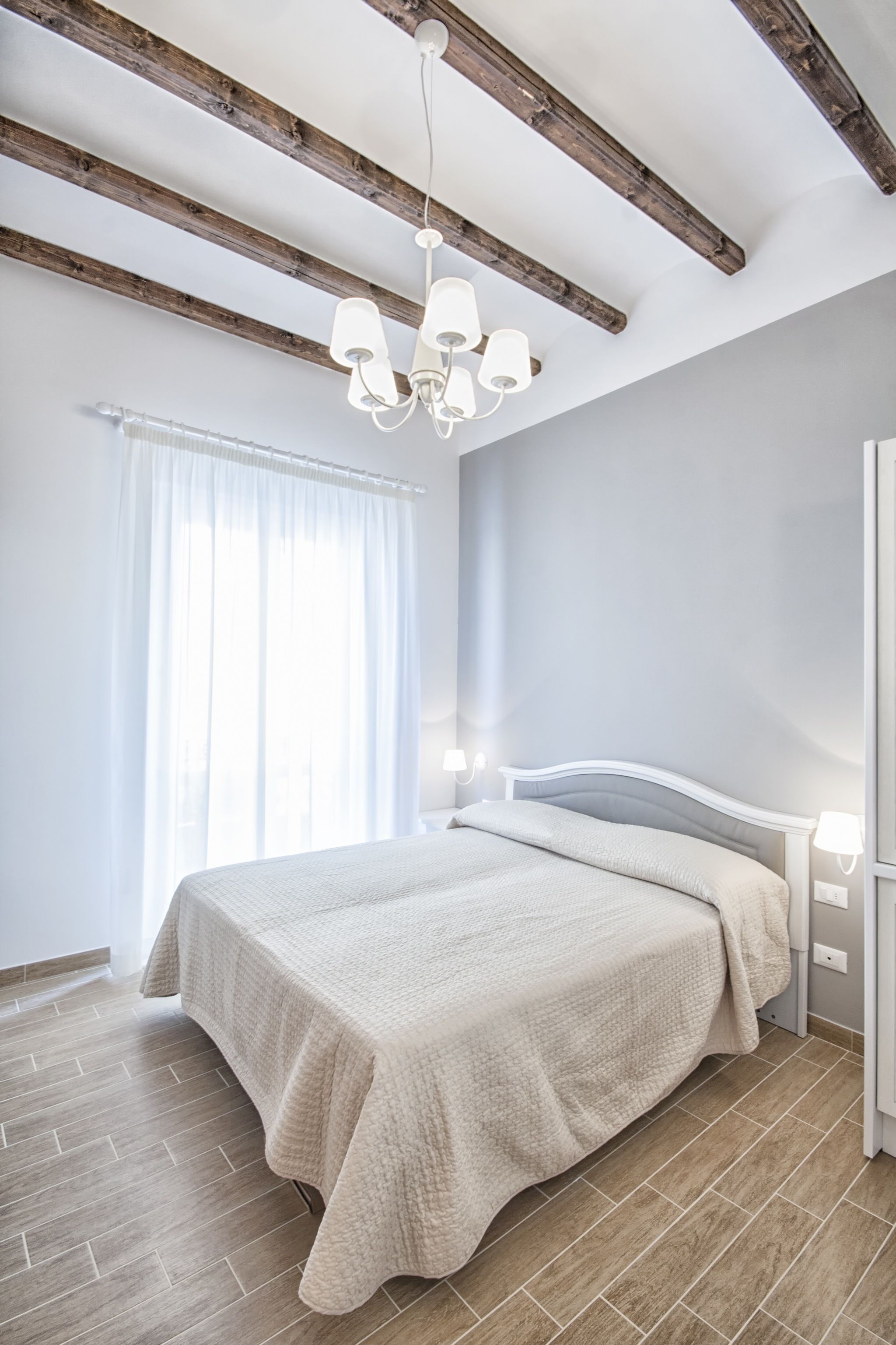 Une chambre à coucher avec un lit, des poutres en bois, un lustre et une fenêtre avec des rideaux.