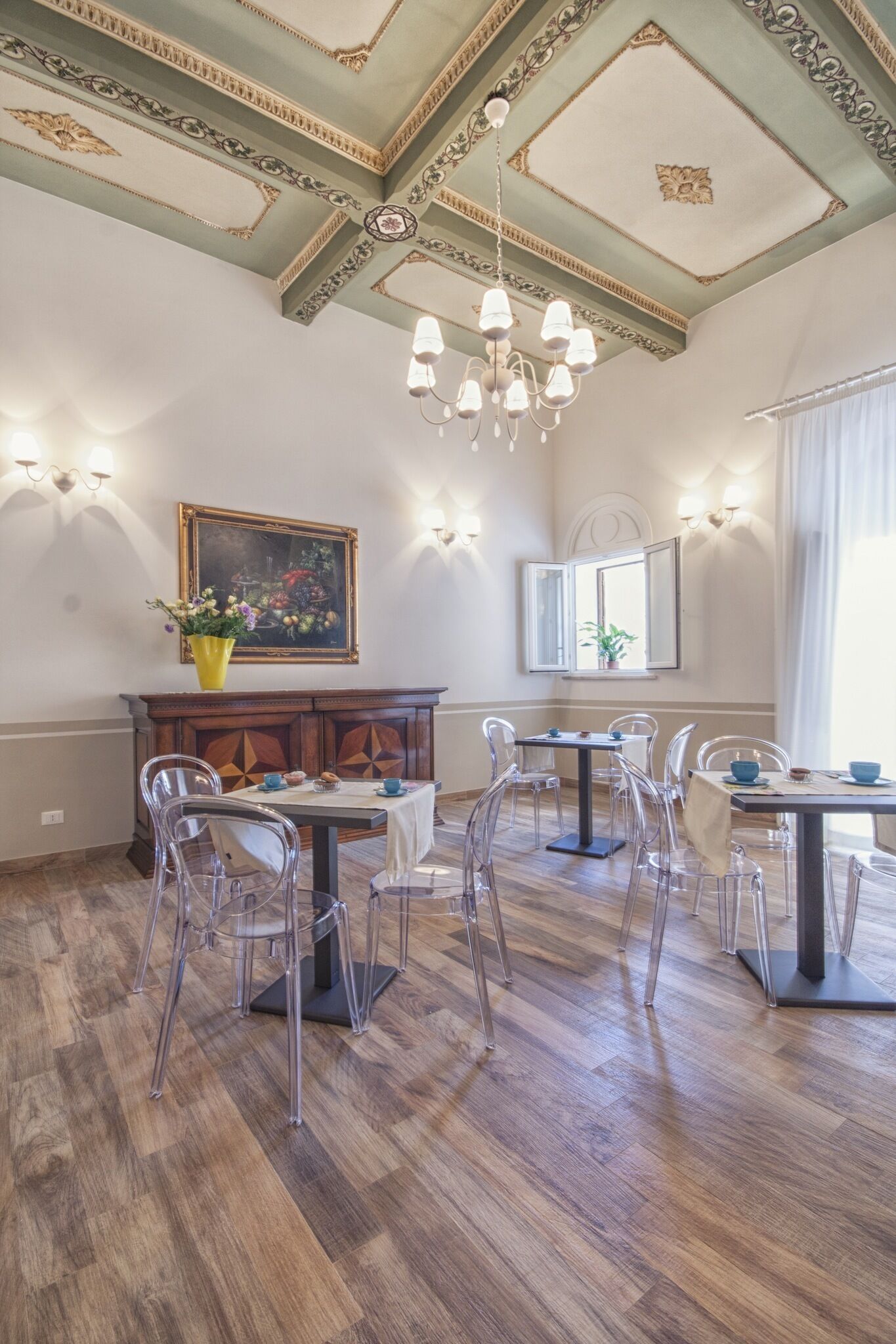 A dining area with wooden flooring, clear chairs, a chandelier, and a painting on the wall.