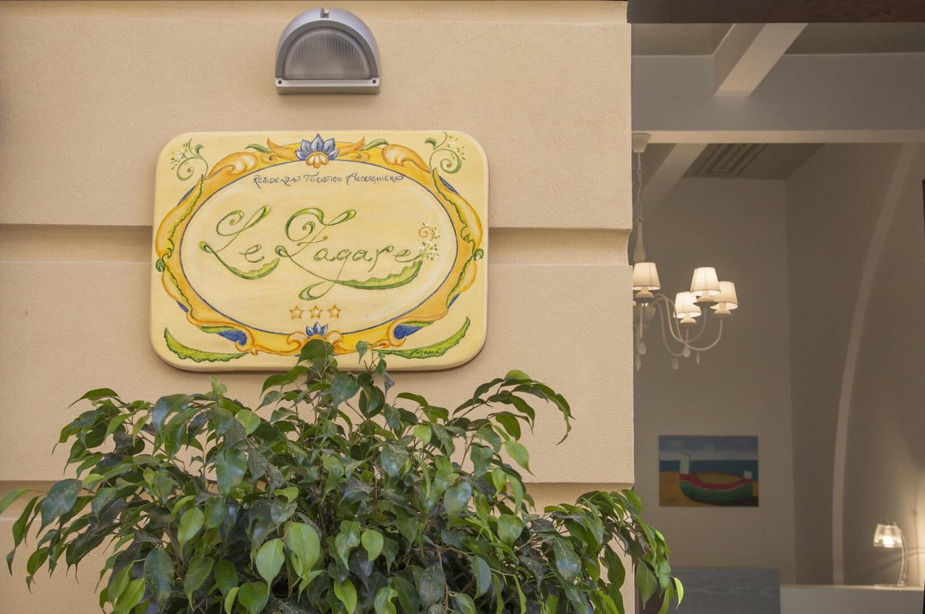 Une enseigne décorative fixée au mur, avec des éléments ornementaux et du texte, un pot de fleurs au premier plan et une vue partielle d’une pièce avec une lampe et une œuvre d’art.