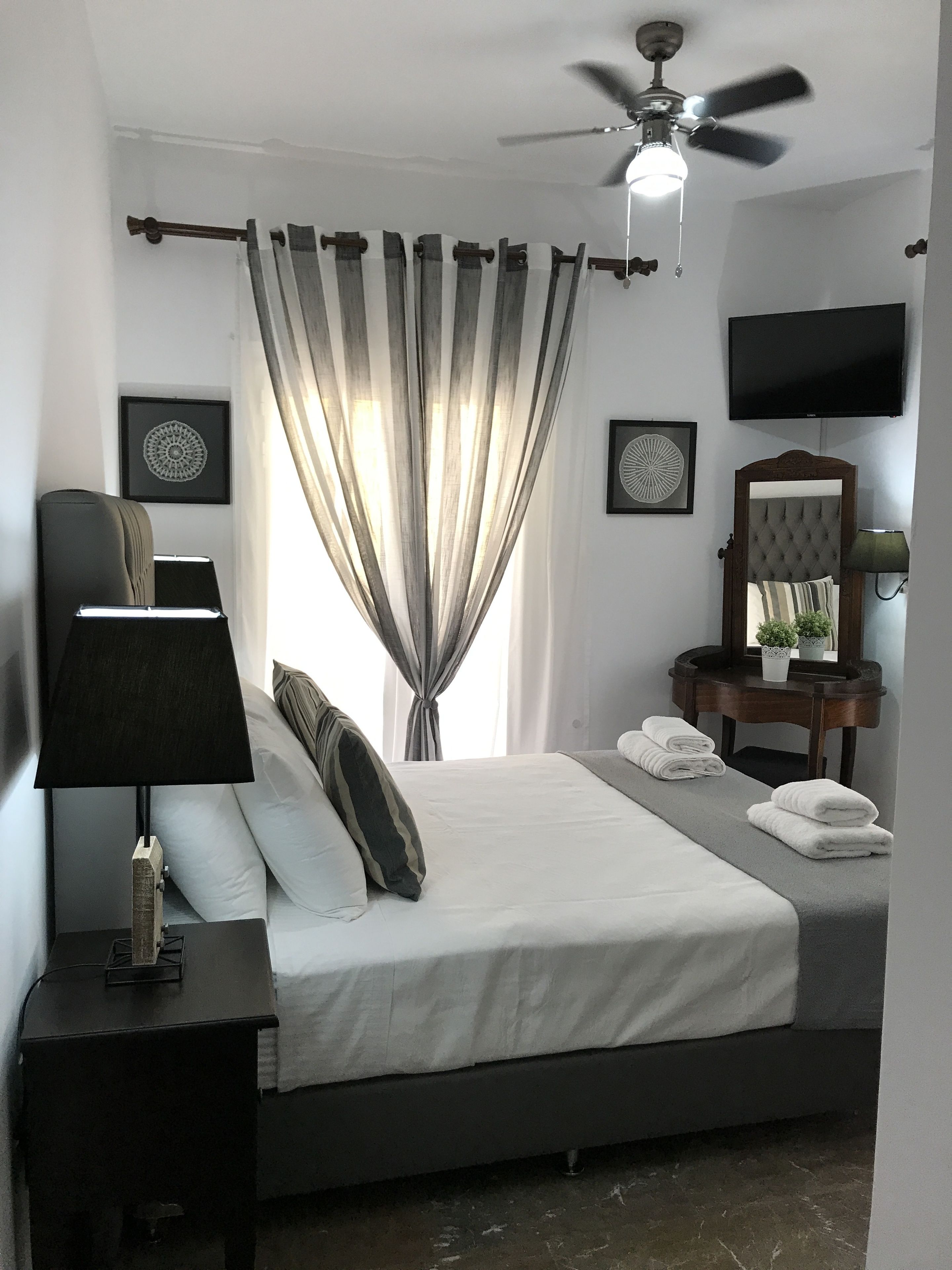 A modern bedroom with a bed, a bedside table with a lamp, a ceiling fan, a TV mounted on the wall, and a window with curtains.
