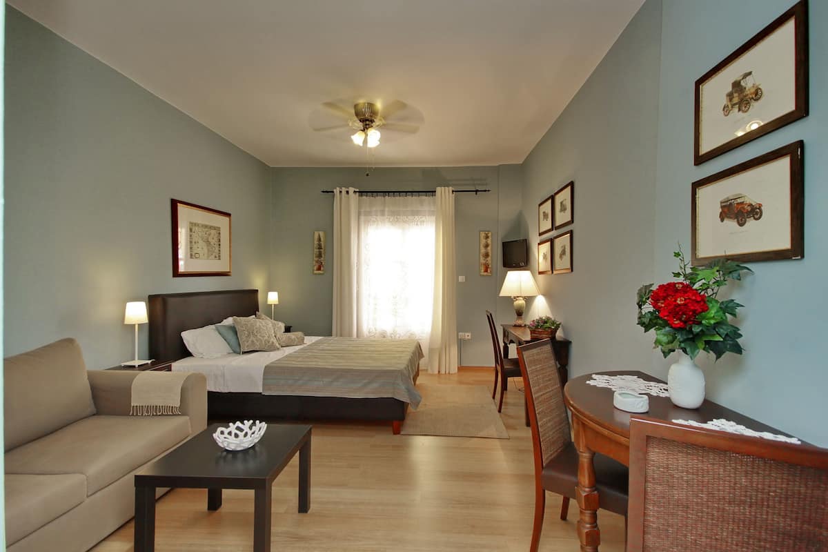 A hotel room with a bed, a sofa, a dining table, and a chair. There is a ceiling fan, a window with curtains, and framed pictures on the wall.