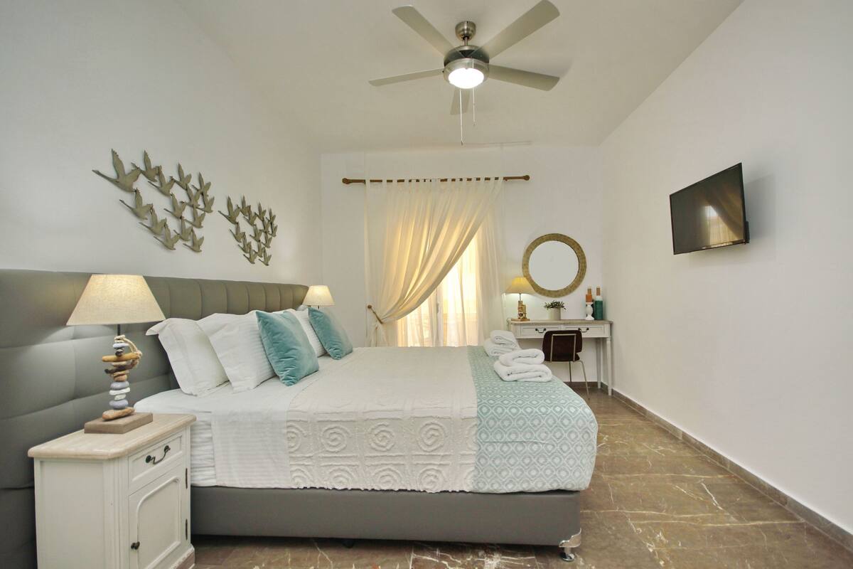 A bedroom with a bed, nightstand, ceiling fan, wall-mounted TV, and a window with curtains.