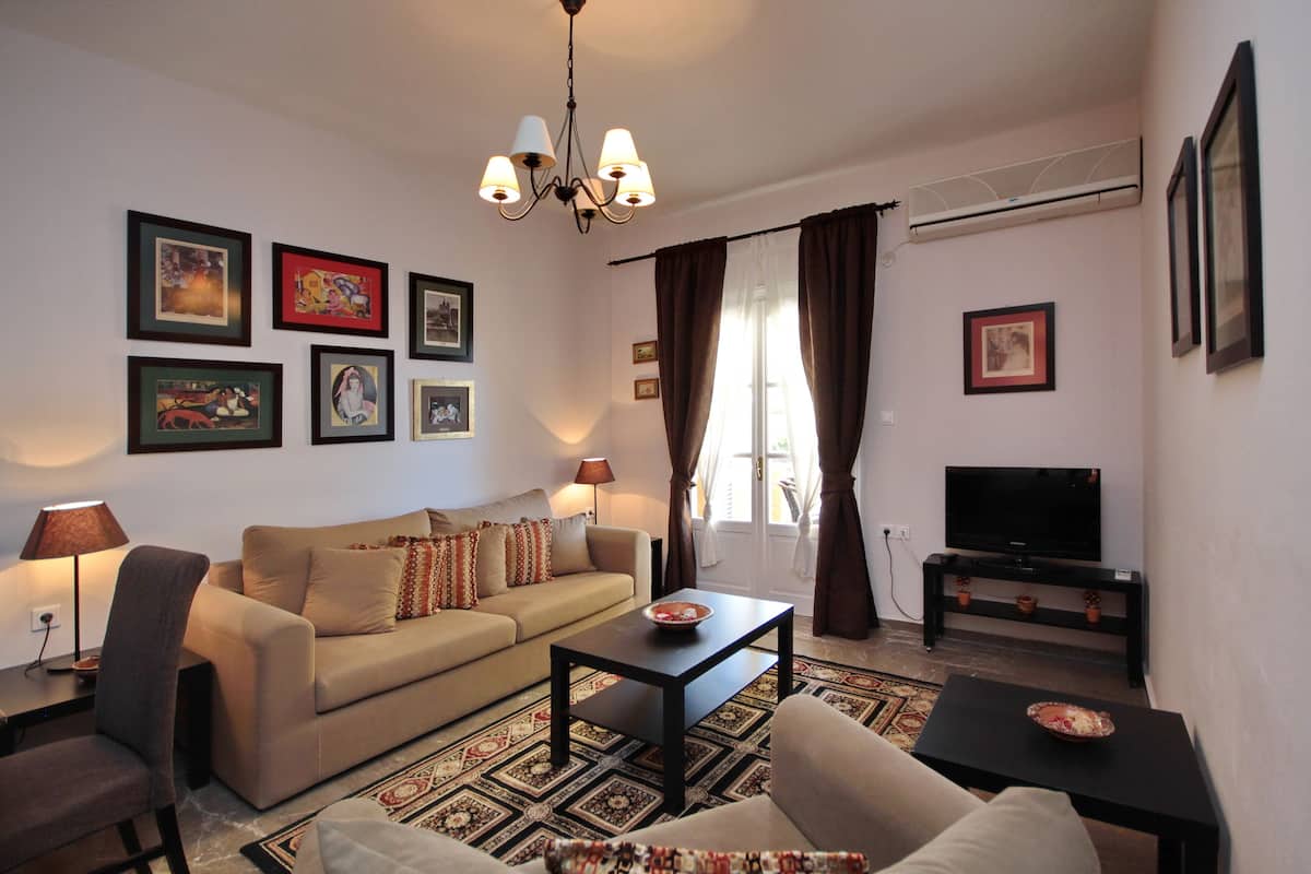 A living room with a sofa, coffee table, and TV stand. There are framed pictures on the wall and a chandelier.
