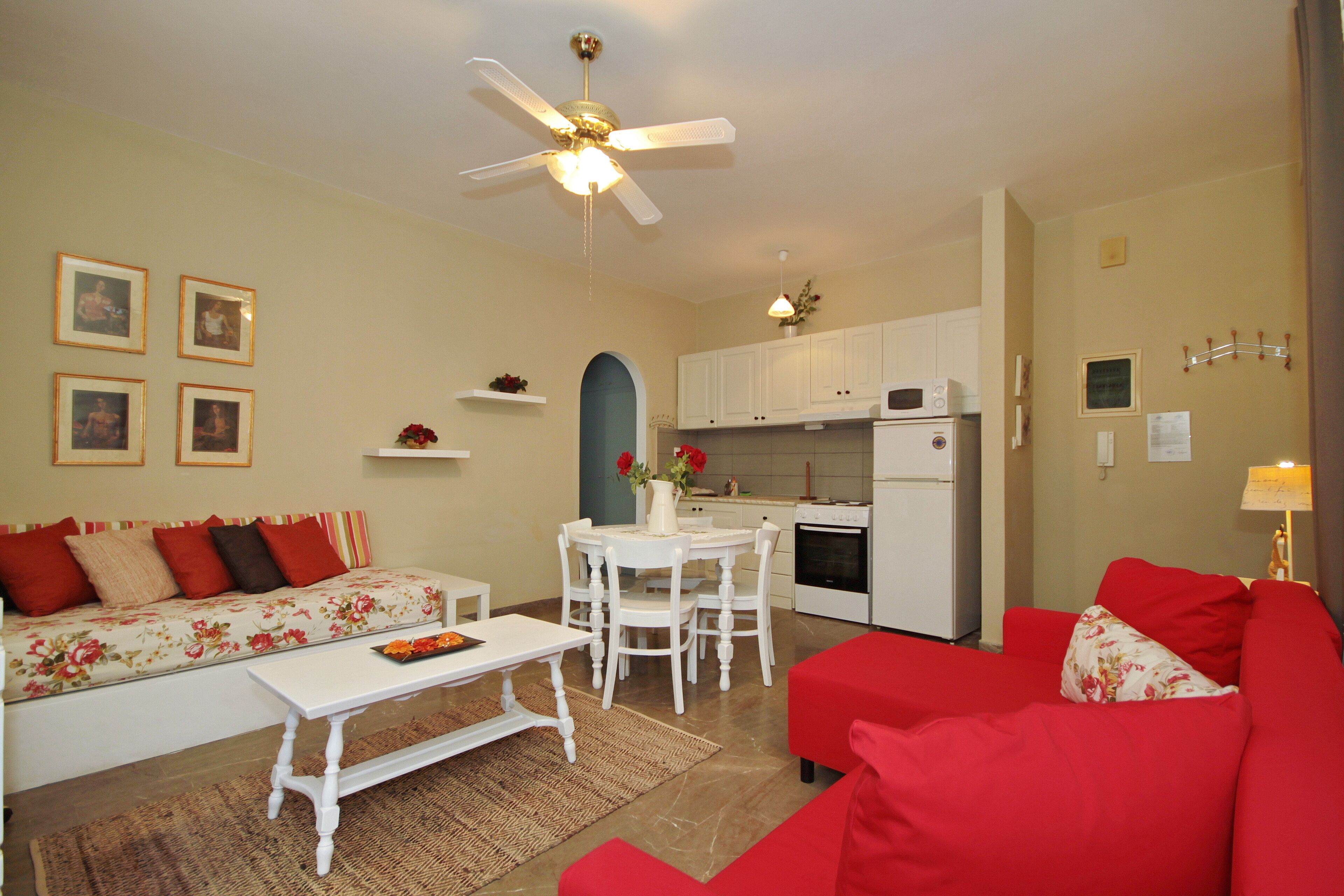 A living room with a red sofa, a white coffee table, and a dining area with a white table and chairs. There is a kitchen with white cabinets and appliances.