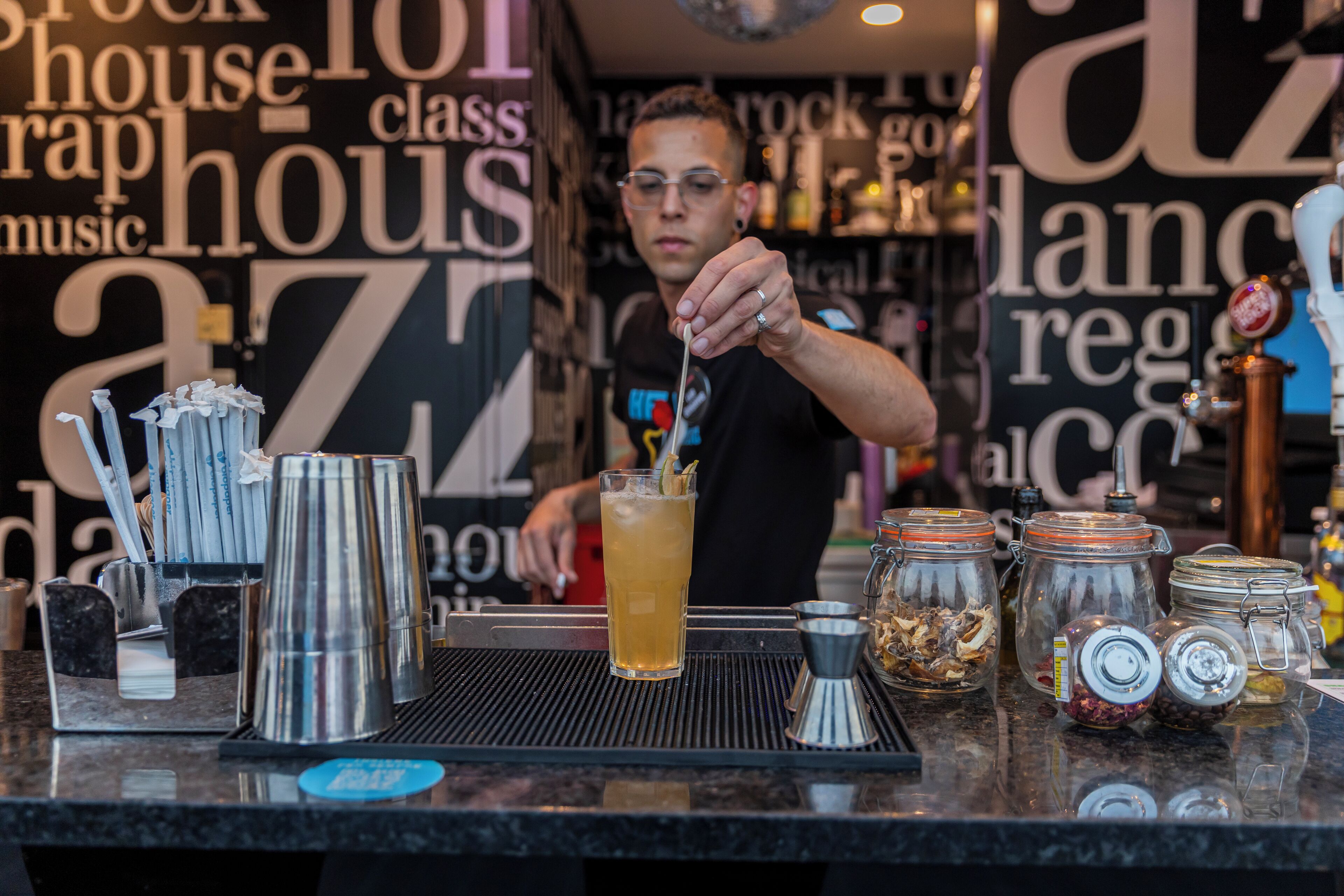 A bar area with a bartender preparing a drink, bar tools, and various containers on the counter.