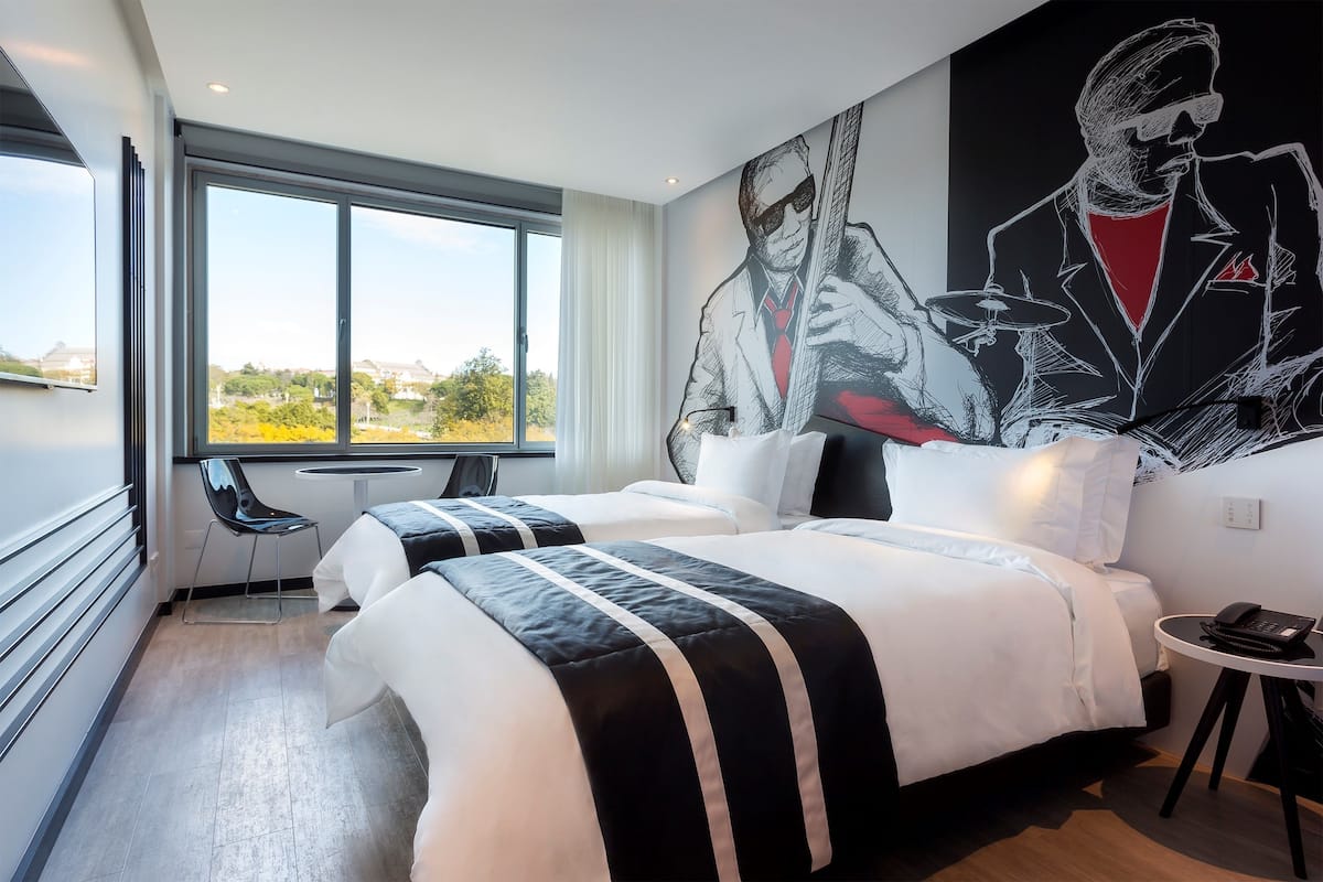 A hotel room with two beds, a large window, and a mural of a man playing drums.