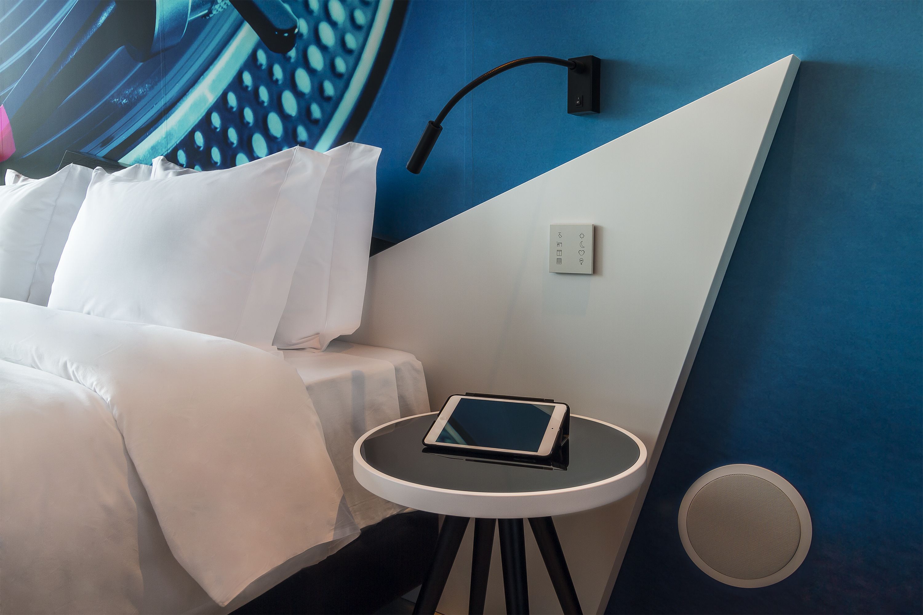 Un lit bien fait, avec du linge de lit blanc, une table de chevet sur laquelle se trouve une tablette, une lampe fixée au mur et une prise de courant.