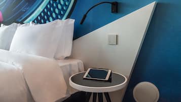 Un lit bien fait, avec du linge de lit blanc, une table de chevet sur laquelle se trouve une tablette, une lampe fixée au mur et une prise de courant.