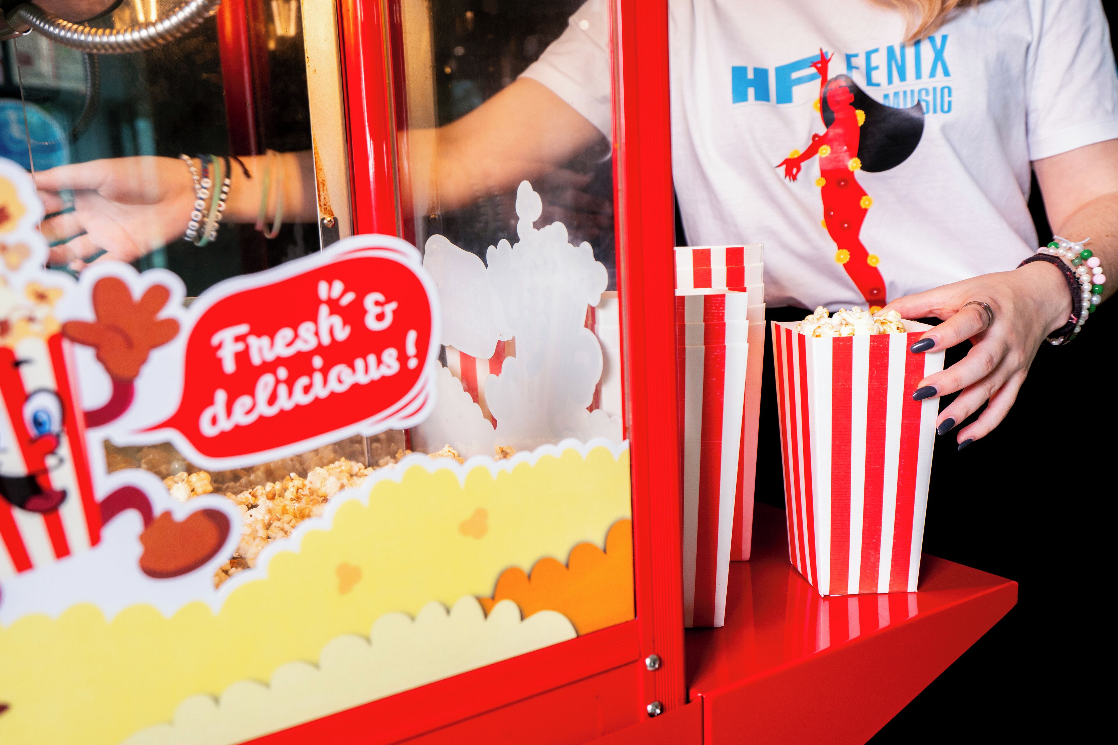A popcorn machine with a sign that reads "Fresh & delicious!" and a person wearing a white shirt with "FENTIX MUSIC" on it.