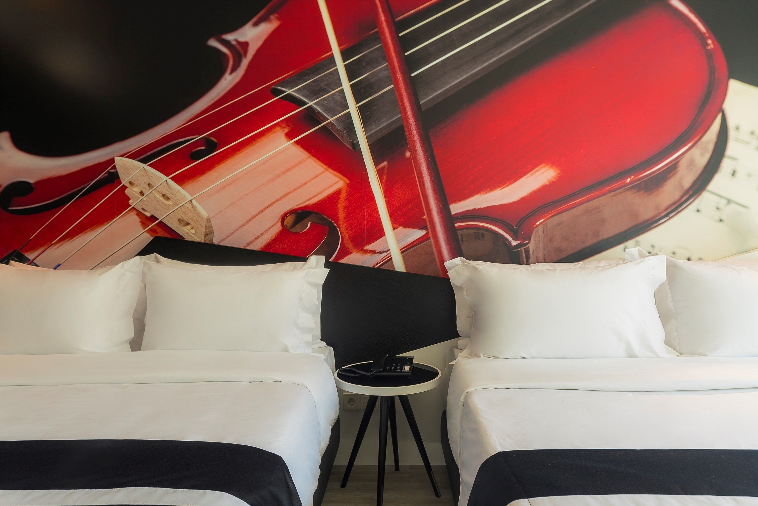 A hotel room with two beds, a small round table, and a large wall mural of a red violin.