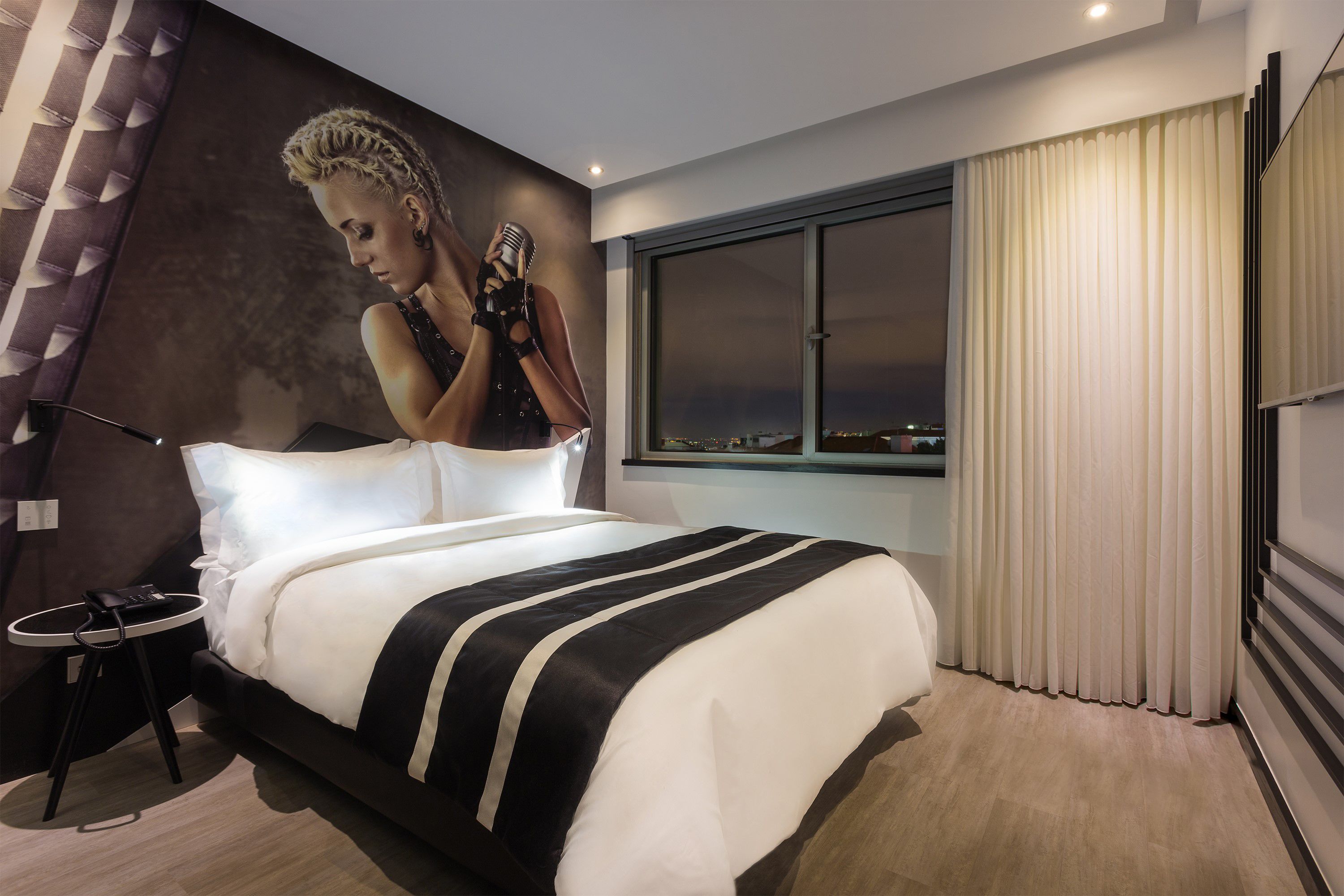 Une chambre d’hôtel moderne avec un grand lit, une petite table de chevet, une fenêtre avec des rideaux et une vue sur la ville.