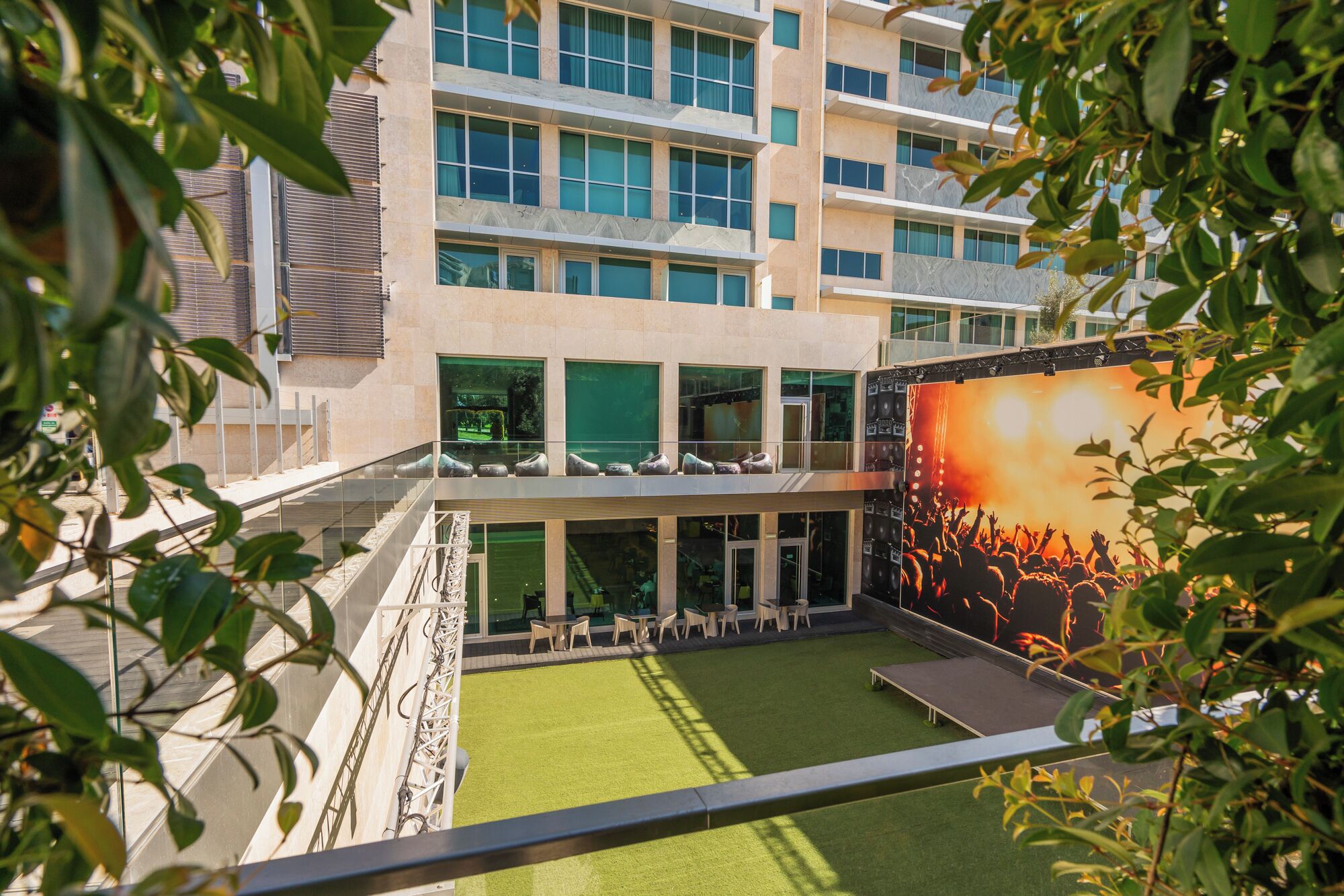 Un edificio moderno con fachada de vidrio, zona de estar al aire libre y una gran pantalla mostrando un concierto.
