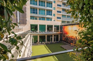 Ένα σύγχρονο κτίριο με γυάλινη πρόσοψη, εξωτερικό χώρο με καθίσματα και μια μεγάλη οθόνη που προβάλλει μια συναυλία.