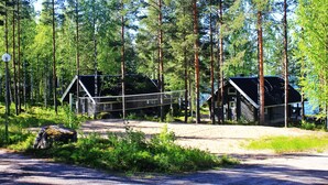 A rustic cabin with a metal bridge connecting to another structure, surrounded by tall pine trees and a dirt path. - Karelia Country Cottages (Kitee)